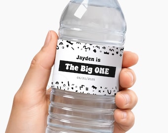 May include: A clear plastic water bottle with a black cap and a white label featuring black abstract shapes. The label text reads: Jayden is The Big ONE 06/21/2025. The bottle has horizontal ridges along the lower section and a smooth upper neck area.