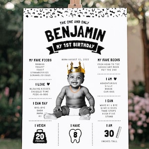 May include: A white birthday milestone sign featuring a black and white photo of a baby wearing a gold crown and chain. The text includes the name Benjamin, My 1st Birthday, birth date, favorite foods, books, and developmental milestones in black.