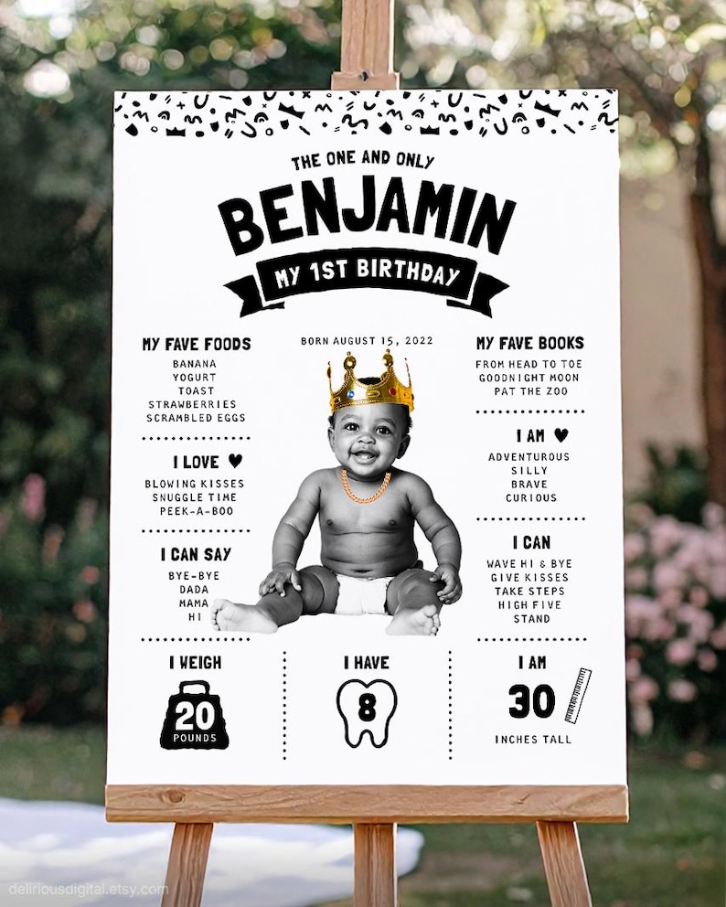 May include: A white birthday milestone sign featuring a black and white photo of a baby wearing a gold crown and chain. The text includes the name Benjamin, My 1st Birthday, birth date, favorite foods, books, and developmental milestones in black.