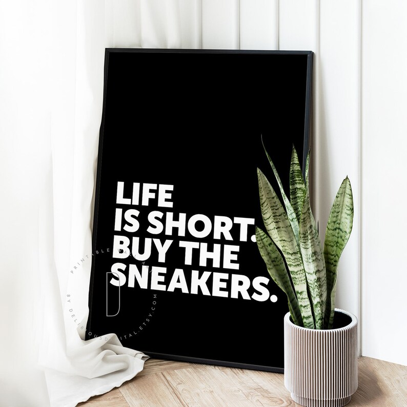 May include: Black and white printable wall art with the text "Life is short. Buy the sneakers."  The art is framed and displayed on a white wall with a green plant in a white pot.