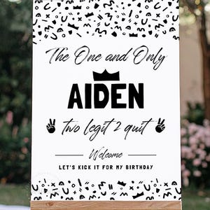 May include: A rectangular white birthday sign featuring black text and abstract shapes. The text reads: The One and Only AIDEN, two legit 2 quit, Welcome, LET'S KICK IT FOR MY BIRTHDAY. A black crown icon sits above the name, with peace sign hand icons.