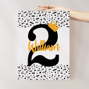 Puede incluir: Un p&oacute;ster de cumplea&ntilde;os imprimible en blanco y negro con un gran n&uacute;mero dos negro con una corona dorada en la parte superior. El nombre William est&aacute; escrito en letras doradas debajo del n&uacute;mero. El fondo es un patr&oacute;n blanco de formas geom&eacute;tricas negras.