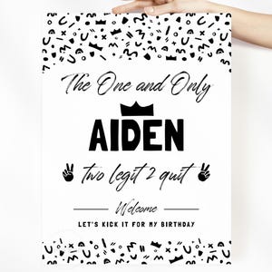 May include: A white rectangular sign with black text and abstract patterns. The text reads: The One and Only AIDEN, two legit 2 quit, Welcome, LET'S KICK IT FOR MY BIRTHDAY. A black crown icon sits above the name, and peace sign icons flank the text.