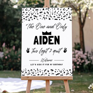 May include: A white rectangular birthday welcome sign on a wooden easel featuring black abstract shapes at the top and bottom, the text The One and Only AIDEN, a crown icon, the text two legit 2 quit with peace sign icons, and Welcome LET'S KICK IT FOR MY BIRTHDAY.