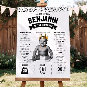 May include: A white birthday milestone sign featuring a black and white photo of a baby wearing a gold crown and chain. The text includes 'THE ONE AND ONLY BENJAMIN MY 1ST BIRTHDAY', 'BORN AUGUST 15, 2022', 'MY FAVE FOODS', 'MY FAVE BOOKS', 'I LOVE', 'I AM', 'I CAN SAY', 'I CAN', 'I WEIGH 20 POUNDS', 'I HAVE 8', and 'I AM 30 INCHES TALL'.