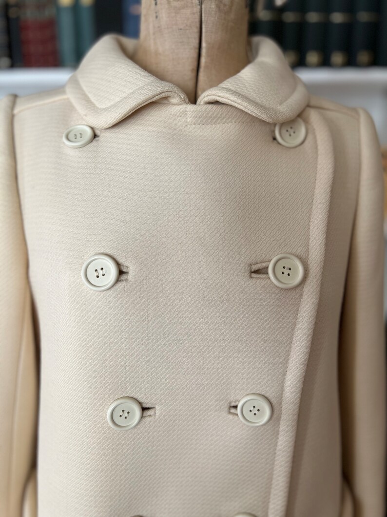 May include: A cream-colored double-breasted coat with a large collar and six white buttons.