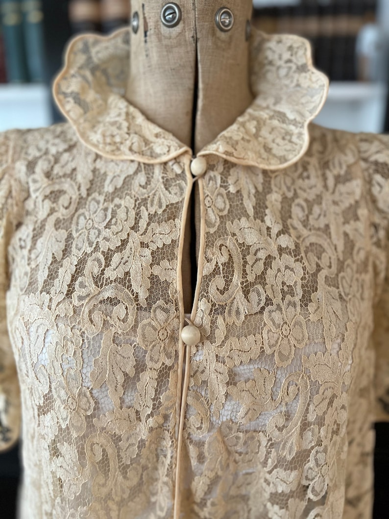 Puede incluir: Una blusa de encaje crema delicada con cuello festoneado y cierre de bot&oacute;n. El encaje presenta un patr&oacute;n floral.