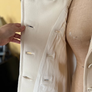 May include: A white double-breasted coat with a button closure. The coat is open and the lining is visible. The lining is also white.