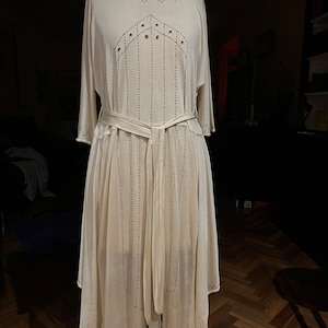 Pode incluir: Um vestido branco fluido com um design geométrico e detalhes de contas pretas. O vestido tem um cinto de laço e uma bainha ligeiramente assimétrica.