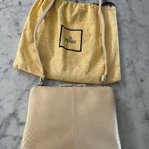 以下が含まれることがあります： クリーム色のヘビ革クラッチバッグ。シルバーの留め具と、それに合わせたショルダーストラップが付いています。バッグには、フェンディのロゴが入った黄色のダストバッグが付いています。