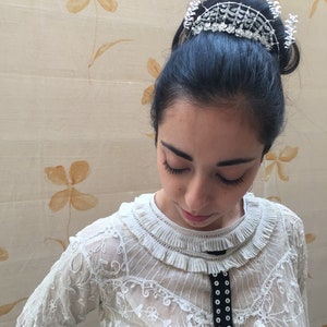 Puede incluir: Una mujer con cabello oscuro lleva una blusa blanca de encaje con una cinta negra y un accesorio para el cabello plateado que parece una corona. El fondo es beige claro con patrones florales marrones.