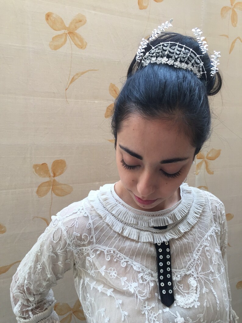 Puede incluir: Una mujer con cabello oscuro lleva una blusa blanca de encaje con una cinta negra y un accesorio para el cabello plateado que parece una corona. El fondo es beige claro con patrones florales marrones.