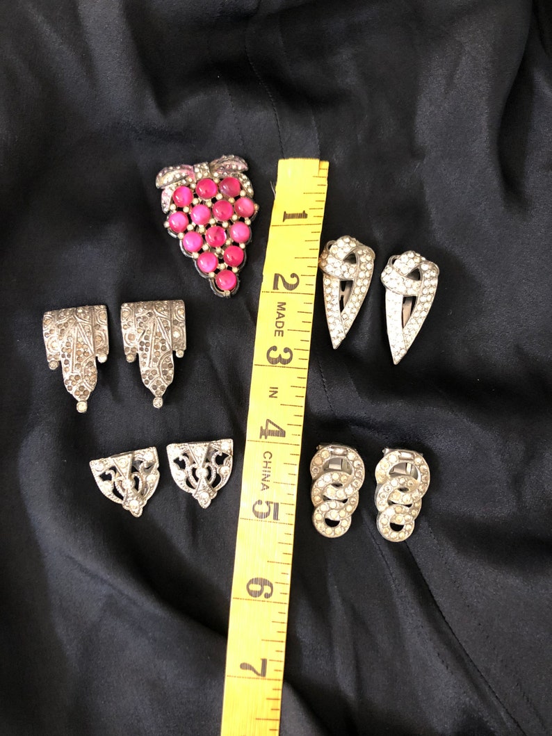 May include: A set of seven vintage silver-toned metal brooches with rhinestones. One brooch features a cluster of pink glass stones in the shape of a grape bunch.  The other brooches are various geometric shapes.