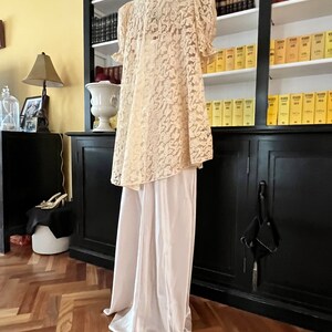Puede incluir: Una blusa blanca de encaje con cuello alto y mangas cortas, combinada con pantalones blancos de sat&eacute;n. El atuendo se muestra en un maniqu&iacute; en una habitaci&oacute;n con una estanter&iacute;a en el fondo.
