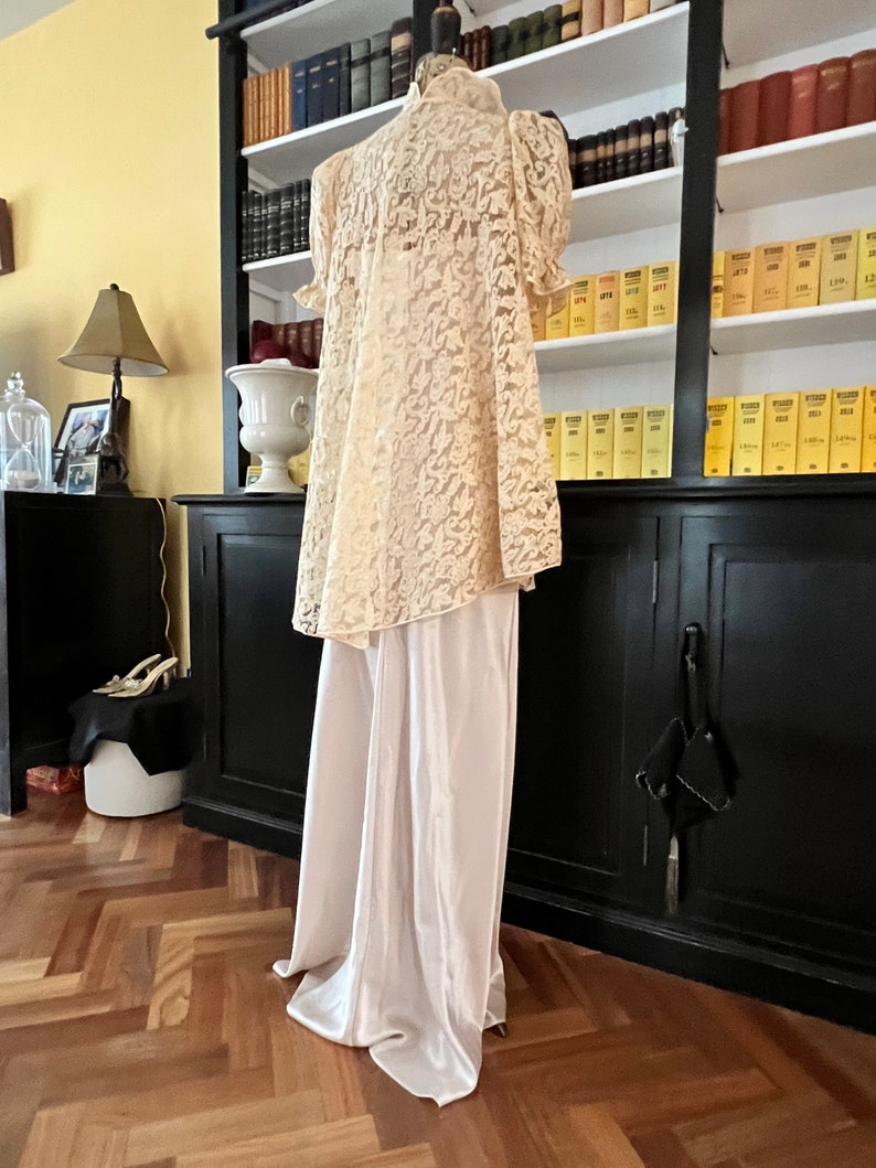 Puede incluir: Una blusa blanca de encaje con cuello alto y mangas cortas, combinada con pantalones blancos de sat&eacute;n. El atuendo se muestra en un maniqu&iacute; en una habitaci&oacute;n con una estanter&iacute;a en el fondo.