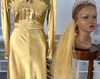 May include: A vintage gold satin gown with long sleeves and a cinched waist, displayed on a mannequin. A pearl and rhinestone brooch adorns the bodice. A mannequin head wears a gold tiara and a matching veil.