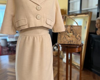 May include: A cream-colored, short-sleeved, cropped jacket with three buttons and a matching skirt. The jacket and skirt are made of a textured fabric. A small, brown, snakeskin handbag is on a wooden stand.