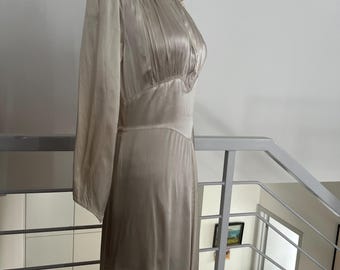 May include: A vintage ivory-colored dress with long sleeves and a high neckline. The dress features a fitted bodice with pleating and a flowing skirt. The dress is displayed on a mannequin, showcasing its elegant design and draping.