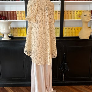 Puede incluir: Un vestido largo de encaje color marfil con un deslizamiento de sat&eacute;n debajo. El vestido tiene un escote alto y un corpi&ntilde;o ajustado con mangas abullonadas. El encaje es un delicado patr&oacute;n floral.