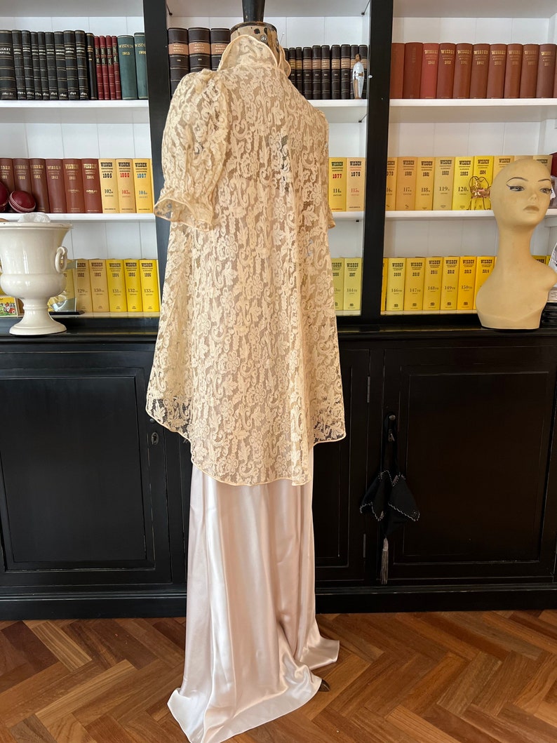 Puede incluir: Un vestido largo de encaje color marfil con un deslizamiento de sat&eacute;n debajo. El vestido tiene un escote alto y un corpi&ntilde;o ajustado con mangas abullonadas. El encaje es un delicado patr&oacute;n floral.