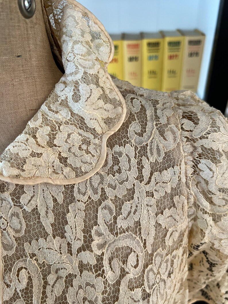 Puede incluir: Una blusa de encaje blanco con cuello festoneado. El encaje es delicado e intrincado, con un patr&oacute;n floral.