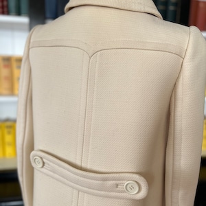 May include: A cream-colored wool coat with a double-breasted closure and a single button on the back. The coat has a classic design and a tailored fit.