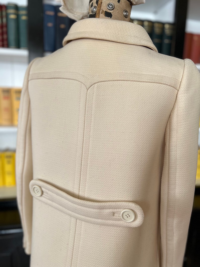 May include: A cream-colored wool coat with a double-breasted closure and a single button on the back. The coat has a classic design and a tailored fit.