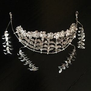 Puede incluir: Una corona floral de plata con pendientes a juego. La corona est&aacute; hecha de delicadas hojas y flores de plata. Los pendientes son largos y colgantes, con un dise&ntilde;o similar al de la corona.
