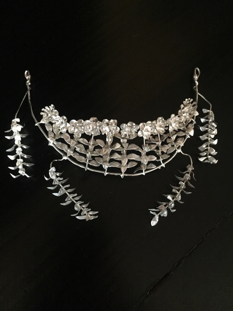 Puede incluir: Una corona floral de plata con pendientes a juego. La corona est&aacute; hecha de delicadas hojas y flores de plata. Los pendientes son largos y colgantes, con un dise&ntilde;o similar al de la corona.