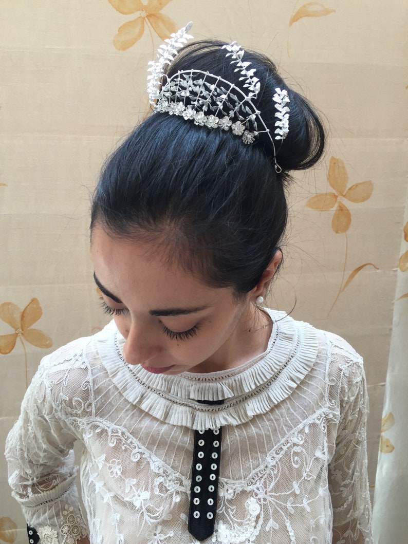 Puede incluir: Una corona blanca con hojas plateadas y pedrer&iacute;a, puesta en el pelo de una mujer.