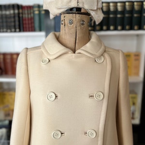 May include: A cream-colored double-breasted coat with a large white bow on the collar. The coat has six buttons down the front.