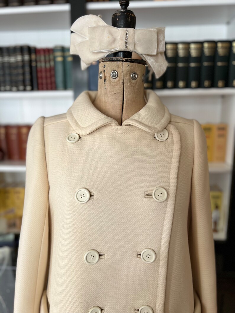 May include: A cream-colored double-breasted coat with a large white bow on the collar. The coat has six buttons down the front.