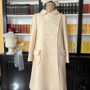 May include: A cream-colored double-breasted coat with large patch pockets. The coat is on a mannequin with a white bow on its head. The mannequin is in front of a bookshelf filled with books.