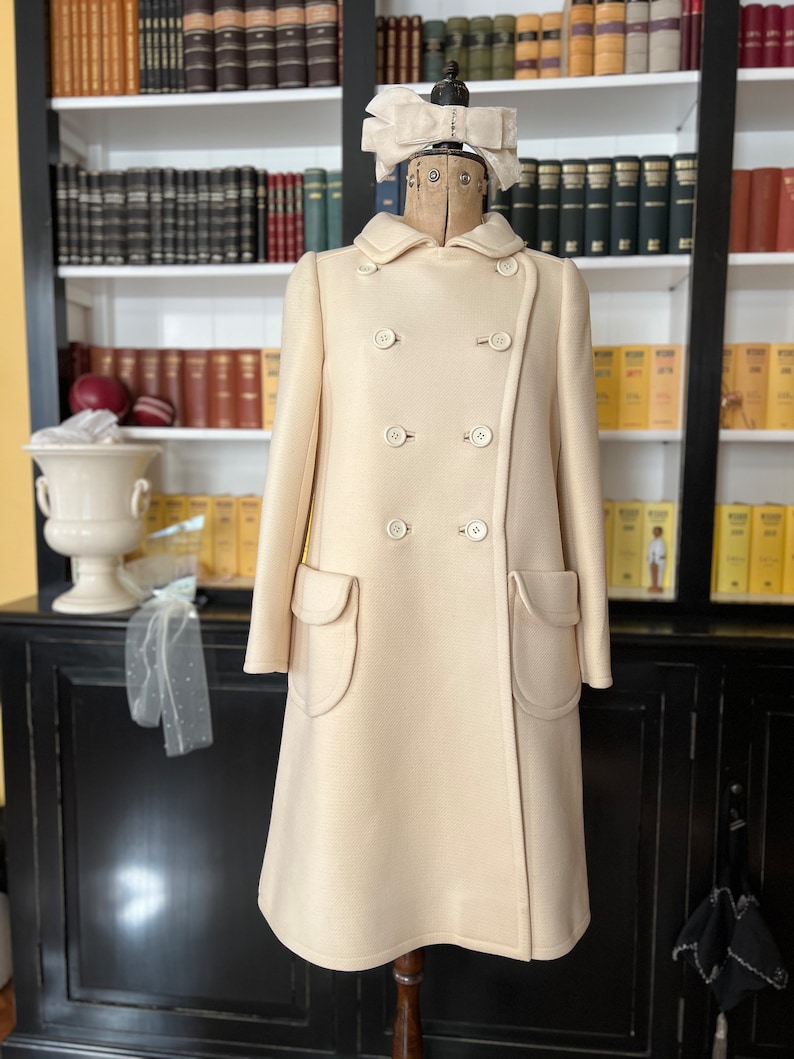 May include: A cream-colored double-breasted coat with large patch pockets. The coat is on a mannequin with a white bow on its head. The mannequin is in front of a bookshelf filled with books.