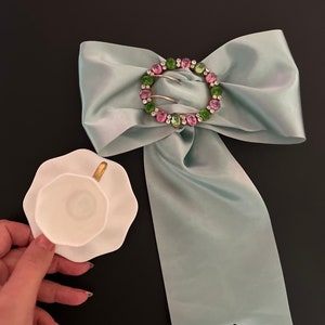 May include: A light blue satin bow with a large, round, silver buckle adorned with pink and green rhinestones. A white teacup and saucer are in the foreground.