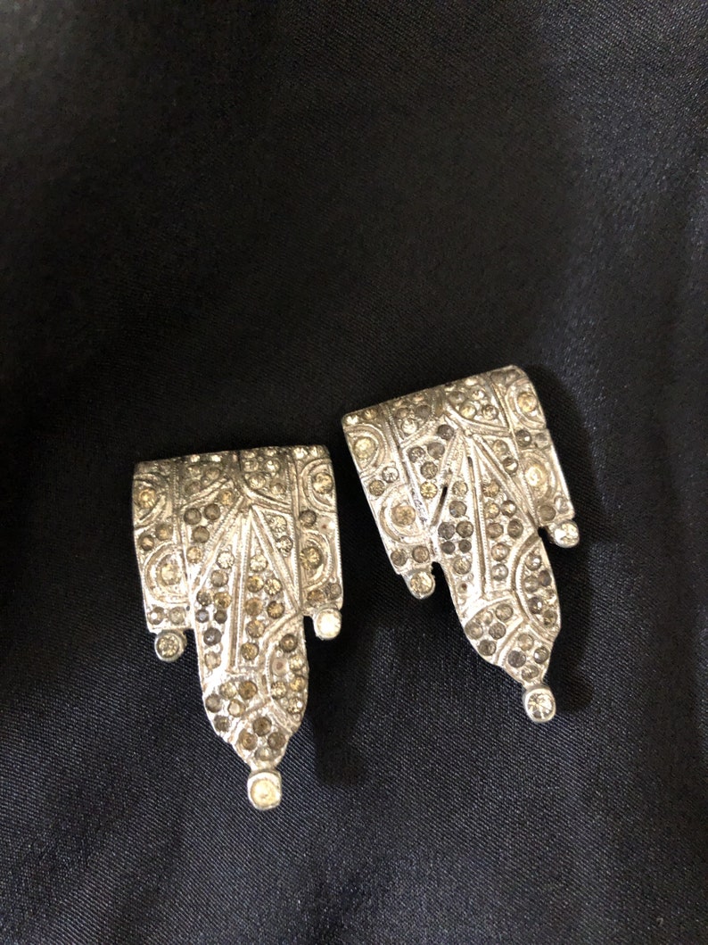 May include: A pair of silver earrings with a geometric design, featuring a cascade of small, clear rhinestones.