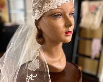 May include: A vintage bridal cap and veil. The cap is crafted from delicate, cream-coloured lace with floral and beaded detailing. The sheer veil is attached to the cap, featuring embroidered floral accents. The cap is displayed on a mannequin head.