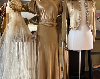 May include: Two mannequins displaying a gold satin dress and a gold satin jacket. The dress has a fitted bodice and a flowing skirt. The jacket has a cropped style with a collar and buttons. Both garments are on display in a room with bookshelves.
