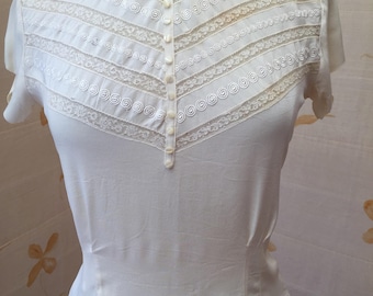 Op de afbeelding: Een wit, kort mouwig blouse met een kant overlay en een rij knopen aan de voorkant.