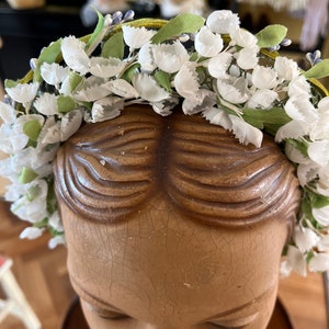 May include: A white flower crown with green leaves and a yellow ribbon. The crown is on a mannequin head.
