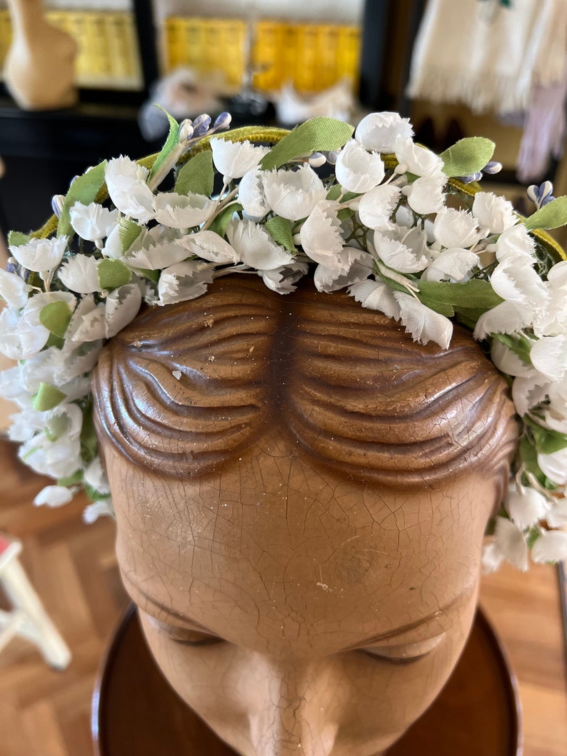 May include: A white flower crown with green leaves and a yellow ribbon. The crown is on a mannequin head.