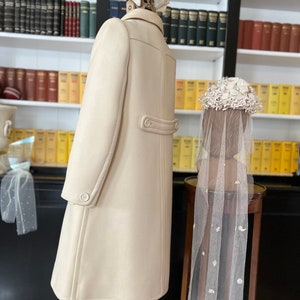 May include: A cream-colored coat with a double-breasted closure and a single button at the back. The coat is displayed on a mannequin with a white flower hat. A white veil with floral embellishments is displayed on a separate mannequin.