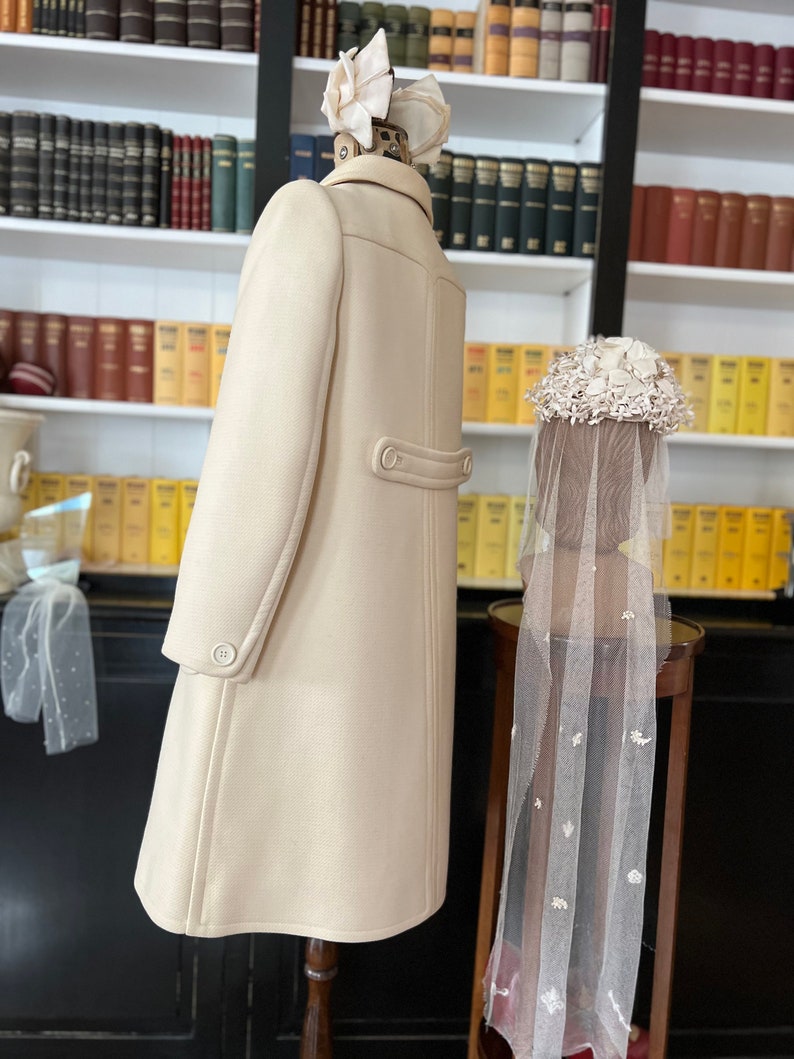 May include: A cream-colored coat with a double-breasted closure and a single button at the back. The coat is displayed on a mannequin with a white flower hat. A white veil with floral embellishments is displayed on a separate mannequin.
