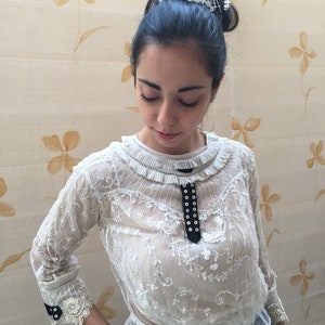 Puede incluir: Una blusa blanca de encaje con una cinta de terciopelo negro y detalle de botones. La blusa tiene un cuello con volantes y mangas 3/4. La modelo lleva una tiara plateada.