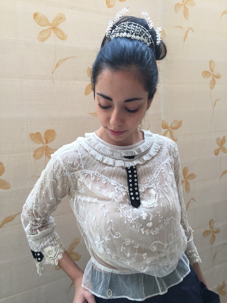 Puede incluir: Una blusa blanca de encaje con una cinta de terciopelo negro y detalle de botones. La blusa tiene un cuello con volantes y mangas 3/4. La modelo lleva una tiara plateada.