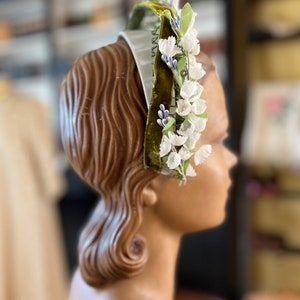 May include: A white headband with a green velvet ribbon and white flowers. The headband is decorated with small blue flowers.