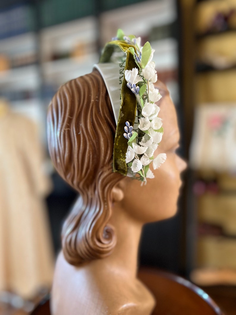 May include: A white headband with a green velvet ribbon and white flowers. The headband is decorated with small blue flowers.