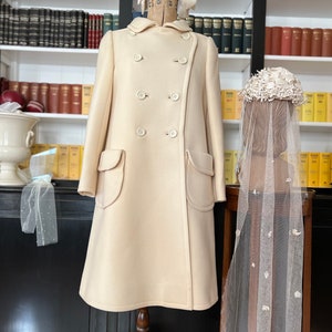 May include: A cream-colored double-breasted coat with two large pockets on the front. The coat is on a wooden mannequin with a white bow on its head. A white veil with floral embellishments is on a separate stand.
