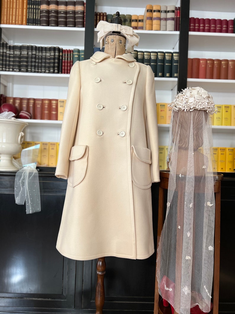 May include: A cream-colored double-breasted coat with two large pockets on the front. The coat is on a wooden mannequin with a white bow on its head. A white veil with floral embellishments is on a separate stand.