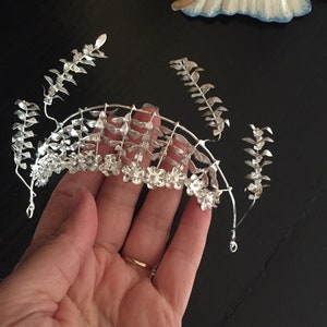 Puede incluir: Corona de plata con dise&ntilde;o floral. La corona est&aacute; hecha de alambre y tiene peque&ntilde;as flores y hojas de plata.